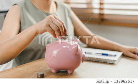 Woman putting coin into pink piggy bank on wooden table at home. Concept of financial planning, money saving for future, retirement investment, and personal budget management with copy space Woman putting coin into pink piggy bank on wooden table at home. Concept of financial planning, money saving for future, retirement investment, and personal budget management with copy space 138962617