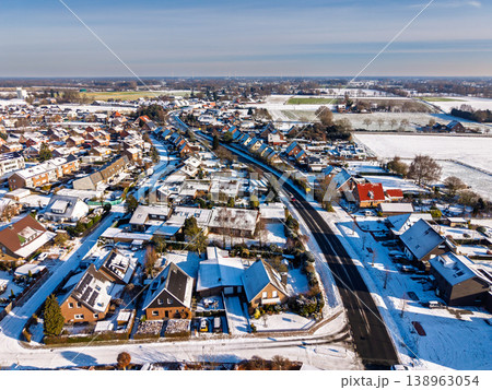 Aerial view of a quiet European town after a snowfall. The image shows snow-covered rooftops, clean asphalt roads, and sunny fields stretching to the horizon on a bright, cold winter morning. 138963054