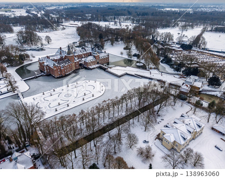 Aerial panorama of a historic moated castle in a snowy winter landscape. Features frozen water, formal geometric gardens covered in white snow, and a majestic brick fortress with towers. 138963060