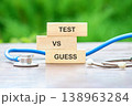 Wooden blocks spelling TEST VS GUESS sit on a brown table, a blue stethoscope nearby. Green foliage blurs in the background, illustrating medical decisions 138963284