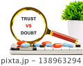 A magnifying glass frames the words TRUST VS DOUBT, with various colorful pills scattered on a white surface, next to a small green plant 138963294