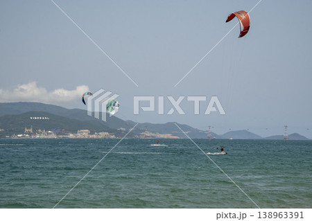 Kitesurfers riding waves on a windy sea with colorful kites, with an amusement park and mountains in the background. Kitesurfers riding waves on a windy sea with colorful kites, with an amusement park and mountains in the background. 138963391