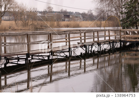 Wooden Pier Bridge over Calm River with Rustic Houses and Reeds in Soft Natural Light Wooden Pier Bridge over Calm River with Rustic Houses and Reeds in Soft Natural Light 138964753