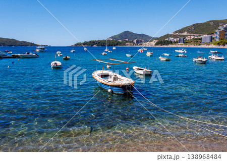 Small boats anchored in crystal clear turquoise water near Adriatic coast with coastal town and mountains in background, sunny summer day, peaceful seaside landscape 138968484