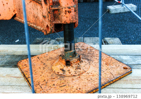 Close up of a rusty crane outrigger support foot on a metal base plate through a temporary safety fence at a roadwork construction site. 138969712