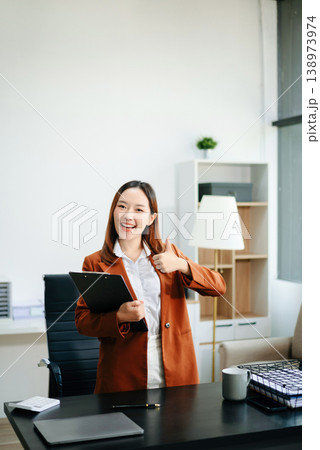 Asian female office worker business suits smiling at camera with working notepad, tablet and laptop documents in office Asian female office worker business suits smiling at camera with working notepad, tablet and laptop documents in office 138973974