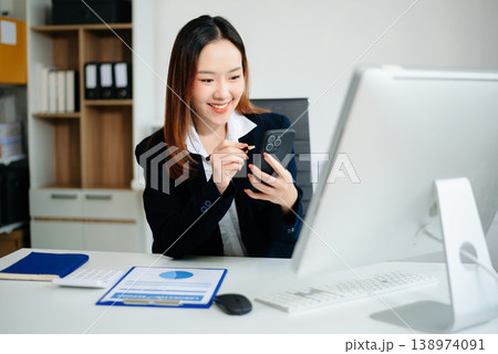 Asian businesswoman in brown blazer working on laptop, analyzing financial data with smartphone. at office Asian businesswoman in brown blazer working on laptop, analyzing financial data with smartphone. at office 138974091