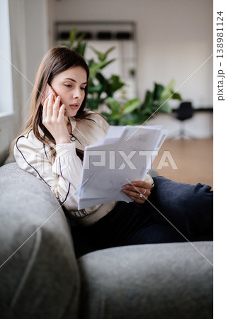 Young woman working from home on a sofa, holding documents and speaking intently on the phone while reviewing paperwork and planning business details during a remote work call Young woman working from home on a sofa, holding documents and speaking intently on the phone while reviewing paperwork and planning business details during a remote work call 138981124