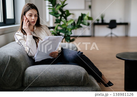 Young businesswoman managing workload from a comfortable grey sofa, holding several documents while engaged in a phone conversation, multitasking efficiently in a modern workspace 138981126