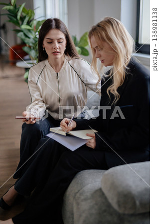 Two young professional women collaborating in a modern office lounge, discussing ideas, taking notes and mentoring each other during a casual coworking meeting Two young professional women collaborating in a modern office lounge, discussing ideas, taking notes and mentoring each other during a casual coworking meeting 138981138