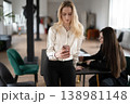 Blonde businesswoman checking her mobile phone, using social media or communicating with clients while standing beside a desk, collaborating with a colleague in a bright office space 138981148