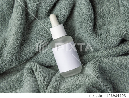 Glass dropper bottle with blank label mockup on sage green bath towel, unbranded cosmetic packaging 138984498