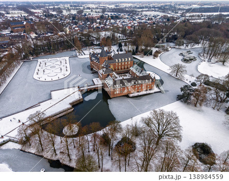Aerial view of Anholt Castle, Wasserburg Anholt, in winter. The historic moated castle and baroque gardens are covered in snow, surrounded by a frozen lake and icy park landscape in Germany. 138984559
