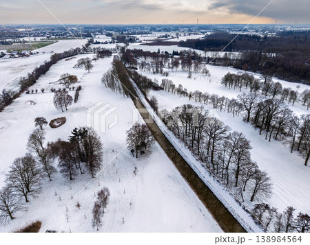 Aerial shot of a snowy golf course and parkland in Germany. A narrow river cuts through the white winter landscape, with bare trees, distant wind turbines, and a soft sunset glow on the horizon. 138984564