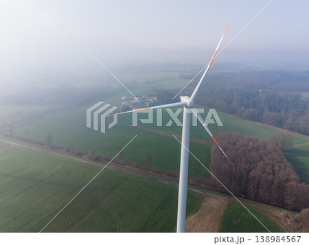 Close-up aerial view of a wind turbine above a misty landscape. Sustainable energy production over green fields, forests, and a biogas plant on a foggy morning in rural Germany. Close-up aerial view of a wind turbine above a misty landscape. Sustainable energy production over green fields, forests, and a biogas plant on a foggy morning in rural Germany. 138984567