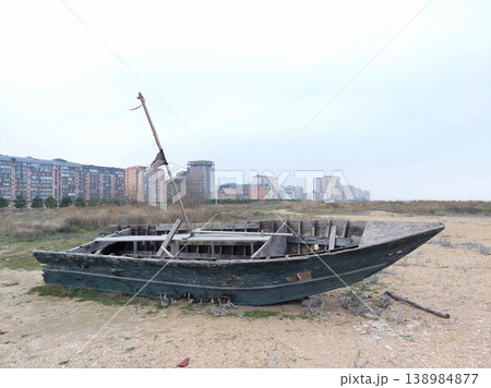 Old abandoned wooden boat on a sandy shore with modern city buildings in the background. 138984877