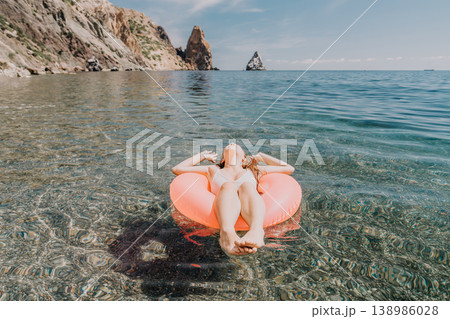 Woman floating sea relaxes on orange inflatable ring in crystal clear ocean water with coastal cliffs in background during peaceful summer vacation Woman floating sea relaxes on orange inflatable ring in crystal clear ocean water with coastal cliffs in background during peaceful summer vacation 138986028