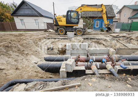 Heavy machinery is digging near a building in a city setting. Workers are installing large pipes for a water system. The ground is dug up and equipment is in use 138986933