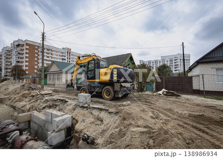 A construction vehicle is digging at a construction site in a neighborhood. Nearby houses and apartment buildings are visible in the background A construction vehicle is digging at a construction site in a neighborhood. Nearby houses and apartment buildings are visible in the background 138986934