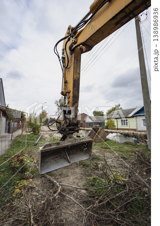 A yellow excavator digs on a construction site in a neighborhood with houses. The sky is cloudy, and some trees are around the area 138986936