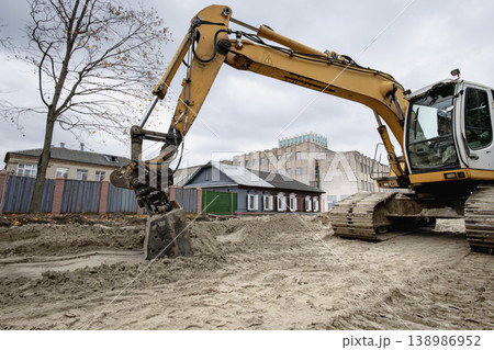 Construction is happening as an excavator digs into the ground near several buildings under a gray sky in an urban area Construction is happening as an excavator digs into the ground near several buildings under a gray sky in an urban area 138986952