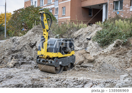 A roller machine is being used in a building site to flatten the ground. Trees with changing leaves are visible nearby. Workers are likely active in the scene A roller machine is being used in a building site to flatten the ground. Trees with changing leaves are visible nearby. Workers are likely active in the scene 138986969