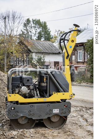 A yellow construction machine sits at a road work site in a small town. The machine is designed for rolling and compacting asphalt or soil A yellow construction machine sits at a road work site in a small town. The machine is designed for rolling and compacting asphalt or soil 138986972