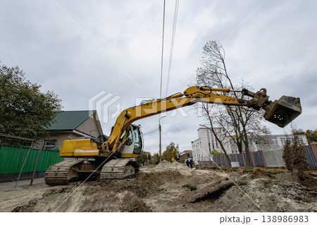 A yellow excavator digs in the ground at a construction site in a residential neighborhood on a cloudy afternoon. Trees and buildings are nearby A yellow excavator digs in the ground at a construction site in a residential neighborhood on a cloudy afternoon. Trees and buildings are nearby 138986983