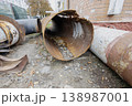 Old and rusty pipes sit on the ground by a brick wall in a city area. Some pipes have holes and dirt inside them. It is afternoon and leaves are scattered around 138987001
