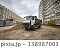 A construction truck is parked on a dirt surface in a residential area. Nearby buildings stand tall as the sky is cloudy and gray 138987003