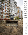 A backhoe loader is on a construction site in front of a tall apartment building. Workers are preparing the ground for a new project in the area 138987005