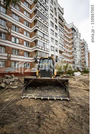A backhoe loader is on a construction site in front of a tall apartment building. Workers are preparing the ground for a new project in the area A backhoe loader is on a construction site in front of a tall apartment building. Workers are preparing the ground for a new project in the area 138987005