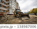 A backhoe is digging and moving earth near a residential building. Workers are focused on the job site as the sky is cloudy. Fences and debris are visible 138987006