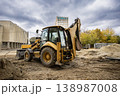 A backhoe loader digs in a construction site located near a tall building. Trees show fall colors against a cloudy sky. Workers may be present 138987008