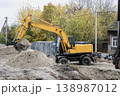 A yellow excavator is digging dirt in a construction site. Trees with autumn leaves are in the background. The site has piles of sand and equipment 138987012