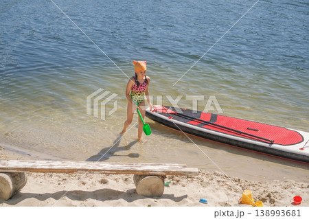 A girl in a swimsuit walks in the water to the shore with a moored board. Sandy beach with a bench by the lake A girl in a swimsuit walks in the water to the shore with a moored board. Sandy beach with a bench by the lake 138993681