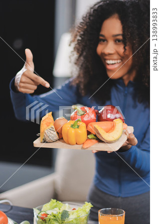 Young nutritionist showing bell peppers during an online video call consulting about healthy eating, sitting at home at a table with vegetables, laptop and measuring tape 138993833