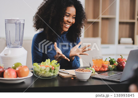 Young nutritionist showing bell peppers during an online video call consulting about healthy eating, sitting at home at a table with vegetables, laptop and measuring tape 138993845