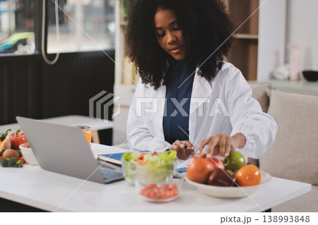 Young nutritionist showing bell peppers during an online video call consulting about healthy eating, sitting at home at a table with vegetables, laptop and measuring tape 138993848