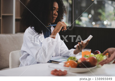 Young nutritionist showing bell peppers during an online video call consulting about healthy eating, sitting at home at a table with vegetables, laptop and measuring tape 138993849
