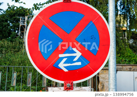 Close up of a red and blue no stopping road sign mounted on a pole beside a stone wall and metal fence. Urban traffic regulation and parking restriction 138994756