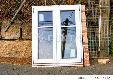 White plastic window frame with glass panes leans against a stone wall beside a road. Discarded building material and renovation debris. White plastic window frame with glass panes leans against a stone wall beside a road. Discarded building material and renovation debris. 138995412