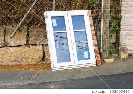 White plastic window frame with glass panes leans against a stone wall beside a road. Discarded building material and renovation debris. White plastic window frame with glass panes leans against a stone wall beside a road. Discarded building material and renovation debris. 138995413