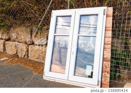 White plastic window frame with glass panes leans against a stone wall beside a road. Discarded building material and renovation debris. White plastic window frame with glass panes leans against a stone wall beside a road. Discarded building material and renovation debris. 138995414