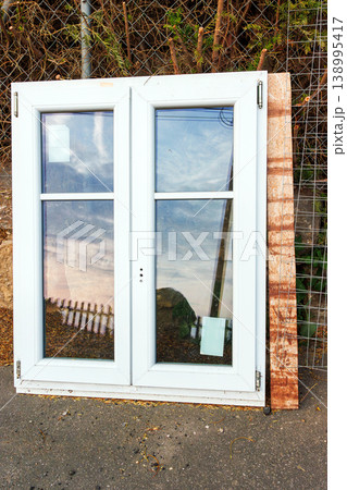 White plastic window frame with glass panes leans against a stone wall beside a road. Discarded building material and renovation debris. White plastic window frame with glass panes leans against a stone wall beside a road. Discarded building material and renovation debris. 138995417