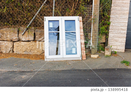 White plastic window frame with glass panes leans against a stone wall beside a road. Discarded building material and renovation debris. White plastic window frame with glass panes leans against a stone wall beside a road. Discarded building material and renovation debris. 138995418