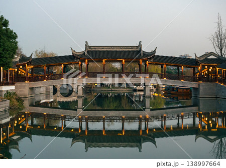 Traditional Chinese covered bridge beautifully illuminated at twilight, its classical architecture reflecting on serene canal water Traditional Chinese covered bridge beautifully illuminated at twilight, its classical architecture reflecting on serene canal water 138997664