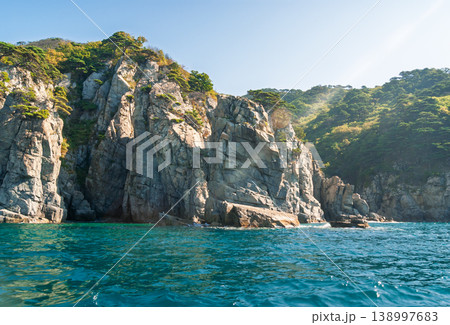 Rugged stone cliffs rise sharply from the deep blue ocean water under a clear, bright summer sky Rugged stone cliffs rise sharply from the deep blue ocean water under a clear, bright summer sky 138997683