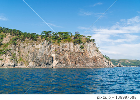 Vast blue sea gently laps against a rugged coastal cliff adorned with green trees under a bright summer sky with wispy clouds during daytime. Vast blue sea gently laps against a rugged coastal cliff adorned with green trees under a bright summer sky with wispy clouds during daytime. 138997688
