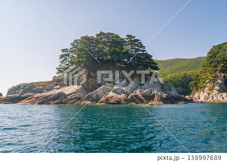 Green pine trees cover a dramatic rocky island shoreline extending into deep blue ocean water on a clear sunny day 138997689